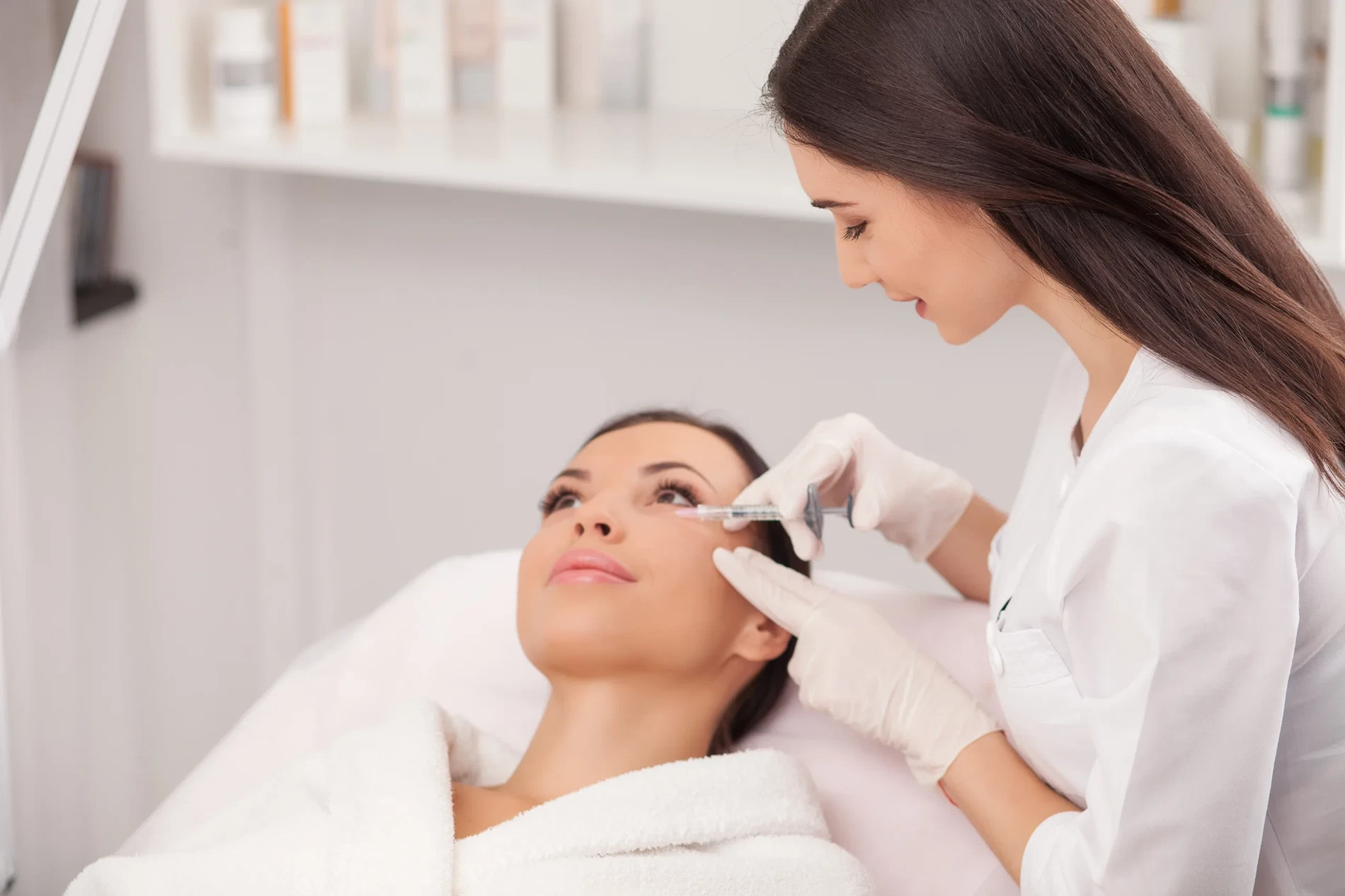 Attractive young woman is getting a collage injection in her face. She is sitting calmly at clinic. The expert beautician is filling female nasolabial wrinkles by hyaluronic acid and smiling