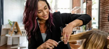 Professional hairstylist with purple-streaked dark hair cutting blonde client's hair with scissors in modern brick-walled salon